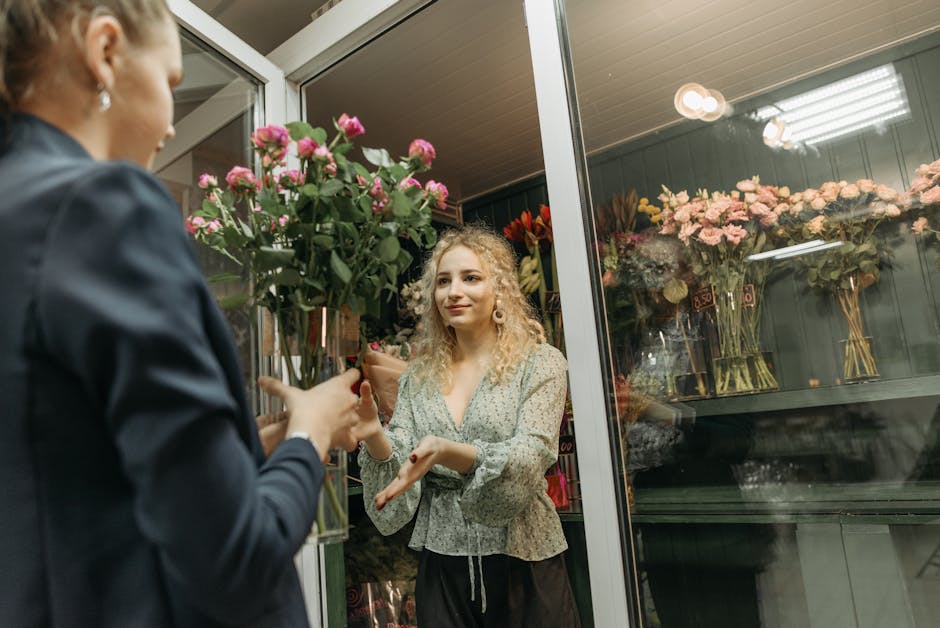 A customer and a florist are exchanging a floral gift inside a well-lit flower shop, viewed through a glass door. The customer, a young woman with curly blonde hair, is smiling softly and holding a bo