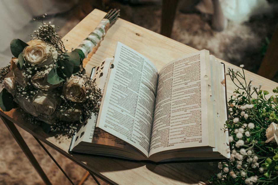 A bouquet of cream-colored roses and small white baby's breath flowers wrapped in white and green striped paper, resting on a wooden table next to an open Bible with densely printed text. The bouquet 