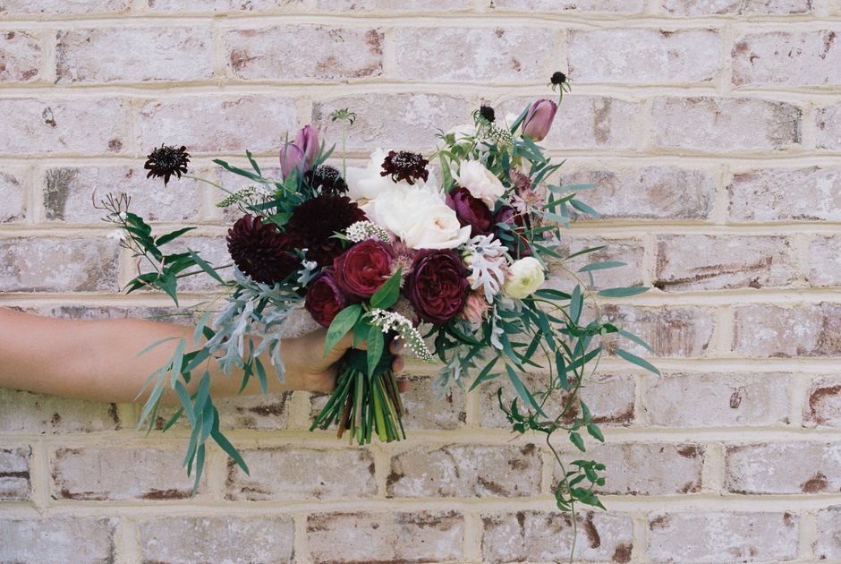 A hand holding a floral bouquet featuring deep burgundy, soft pink, and white roses, complemented by dark purple dahlias, white astilbe, and sprigs of eucalyptus with slender green leaves. The bouquet