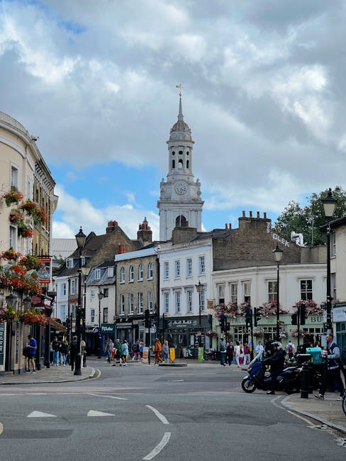 A lively street scene in Tottenham with several boutique shops and cafes lining the two sides of the street, adorned with hanging flower baskets and colorful window displays. The foreground shows pede