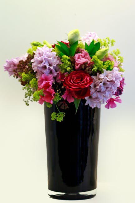 A vibrant bouquet of fresh flowers arranged in a tall, elegant black glass vase, featuring a mix of pink, red, and white blooms. Prominent in the arrangement are a large red rose, soft pink hyacinths,
